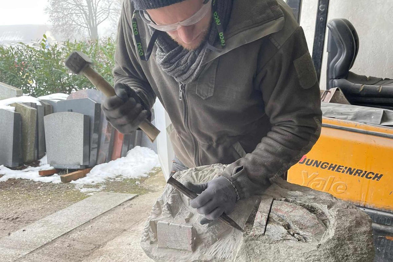 Ein Mann bearbeitet einen Stein mit Hammer und Meißel, im Hintergrund Grabsteine.
