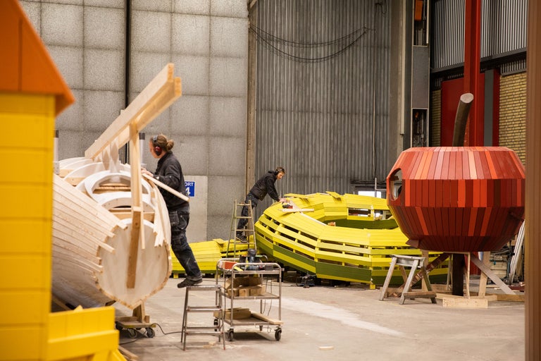 Dans un atelier, une maison en forme de pomme, un tunnel jaune et une autre maison en bois sont en construction.
