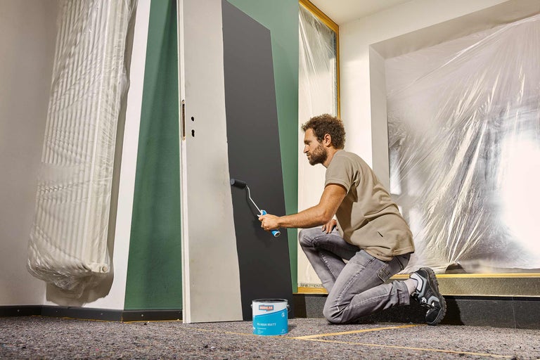 Un homme peint une porte au rouleau dans une pièce recouverte de film plastique, pot de peinture prêt.