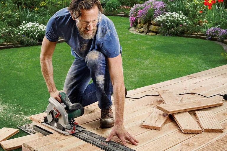 Un homme travaille avec une scie circulaire à main sur une terrasse en bois dans le jardin.