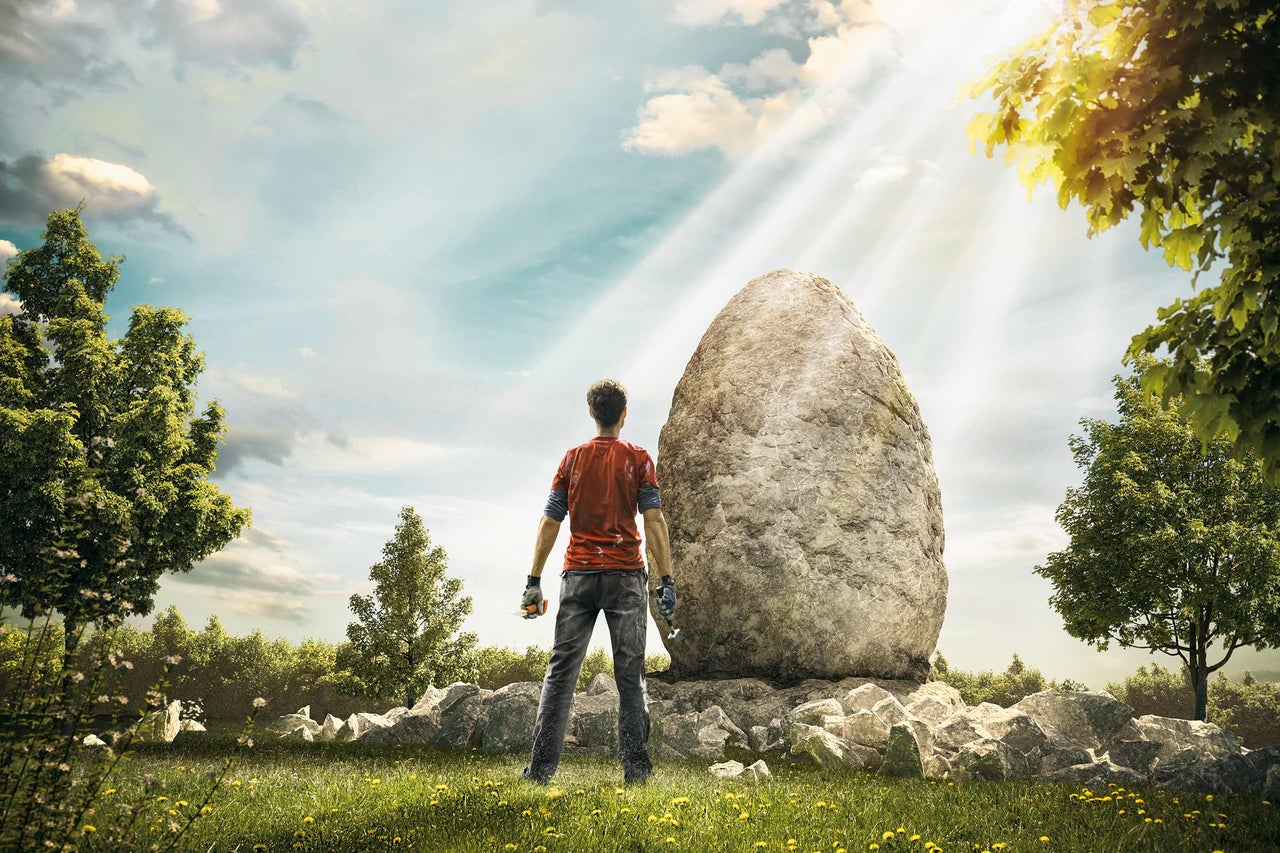 Mann steht vor großem Stein auf einer Wiese mit Bäumen