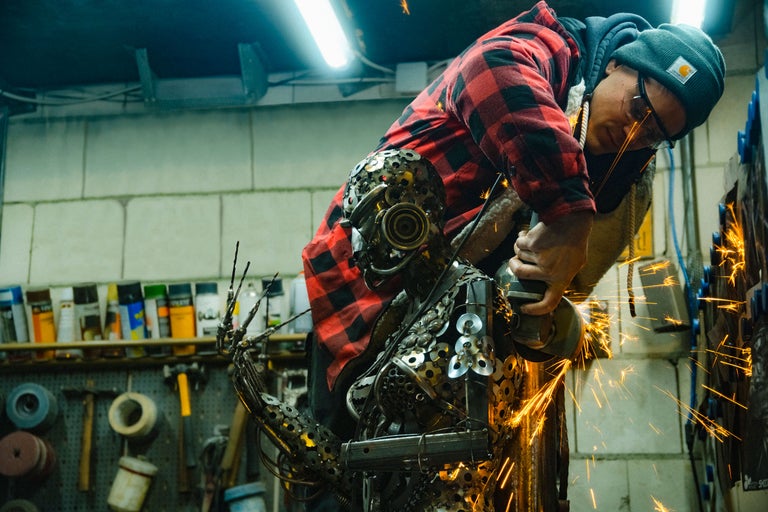 Une personne travaille une sculpture métallique avec une meuleuse d'angle, des étincelles jaillissent.