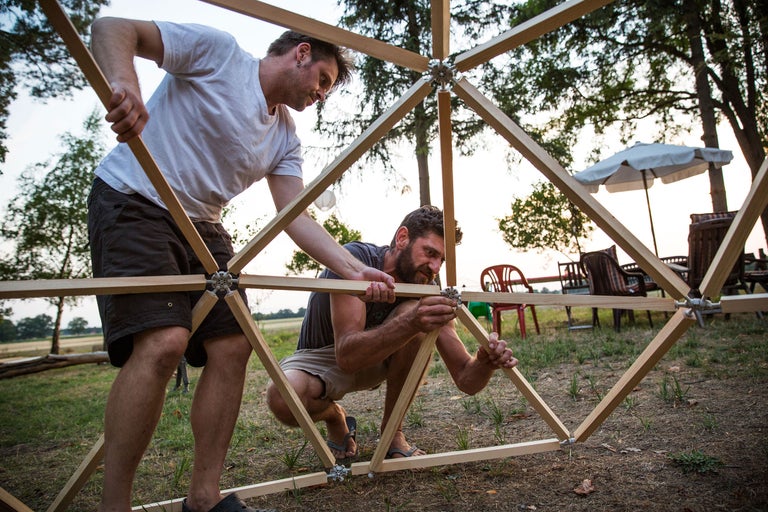 Zwei Personen bauen eine geodätische Kuppel aus Holzstreben und Verbindungselementen im Freien.