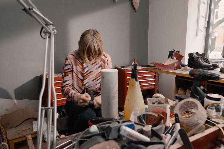 Eine Frau arbeitet in ihrer Werkstatt an der Schuhherstellung, Werkzeuge und Materialien liegen auf dem Tisch.