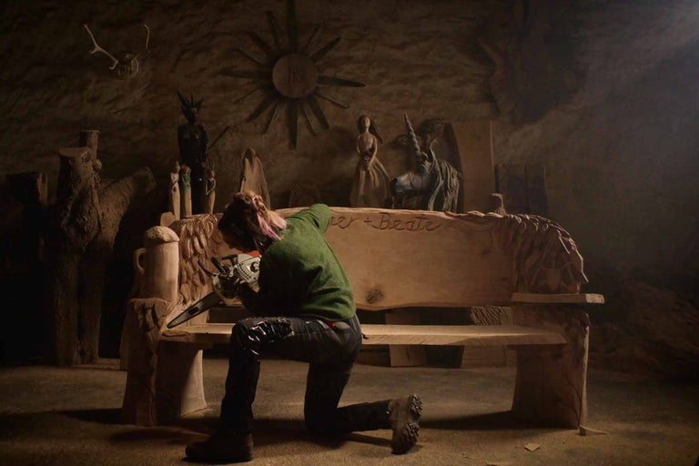 Eine Frau kniet vor einer Holzbank und arbeitet mit einer Kettensäge an ihr. Im Hintergrund stehen verschiedene Holzskulpturen.