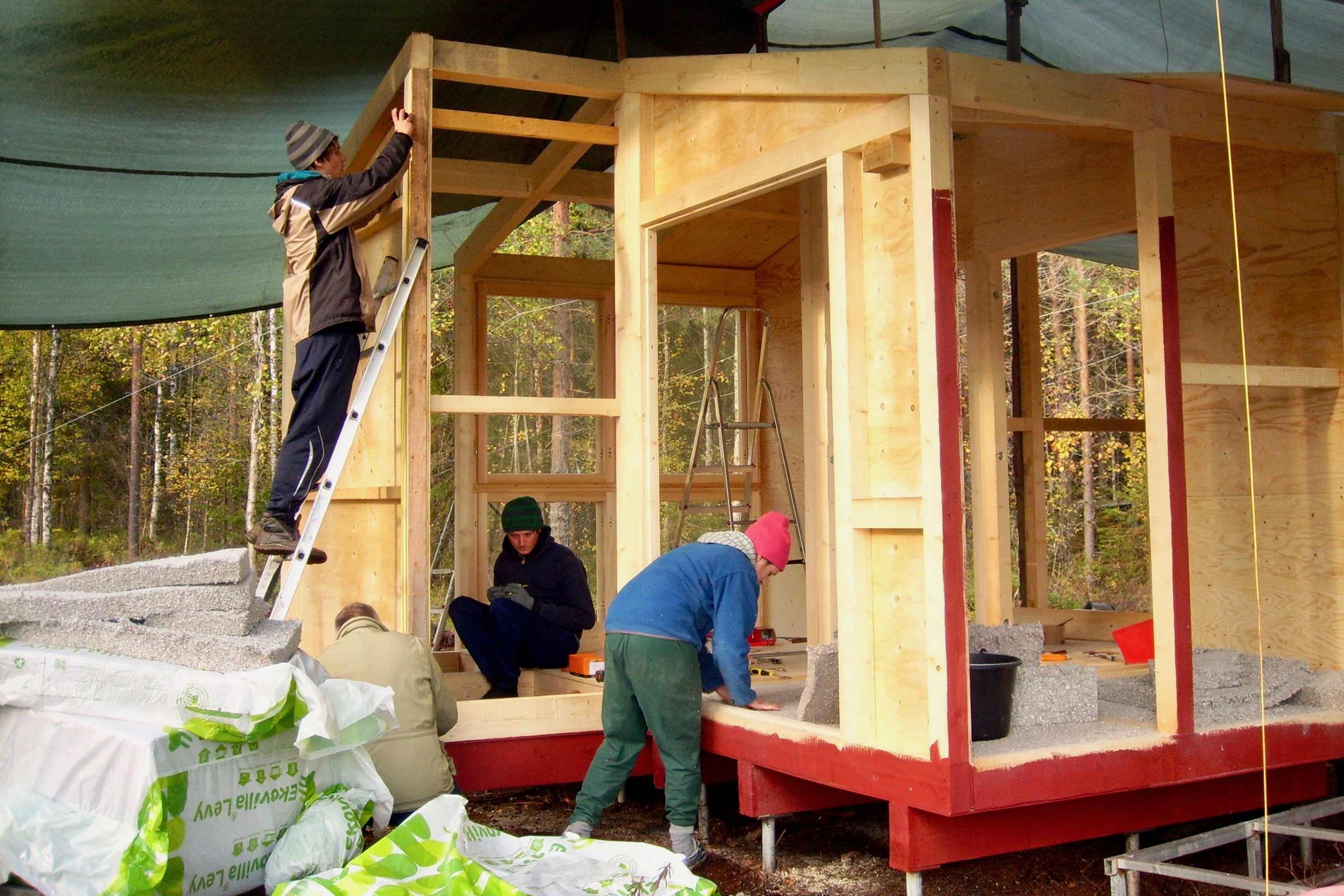 Plusieurs personnes construisent une maison en bois avec une ossature en bois et des panneaux isolants. Le chantier est situé à l'extérieur.