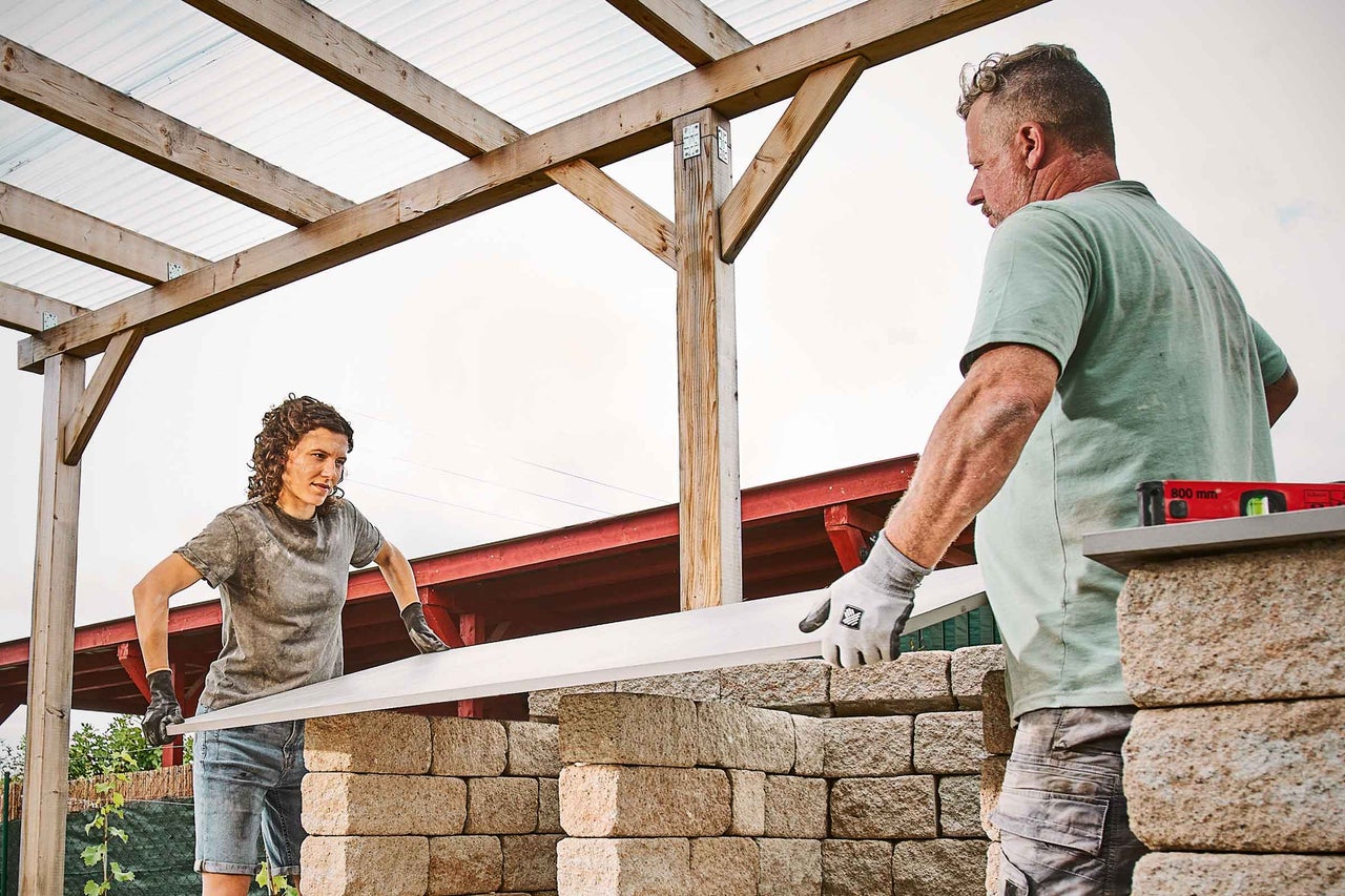 Deux personnes construisent un mur de jardin en blocs de béton sous un abri en bois.
