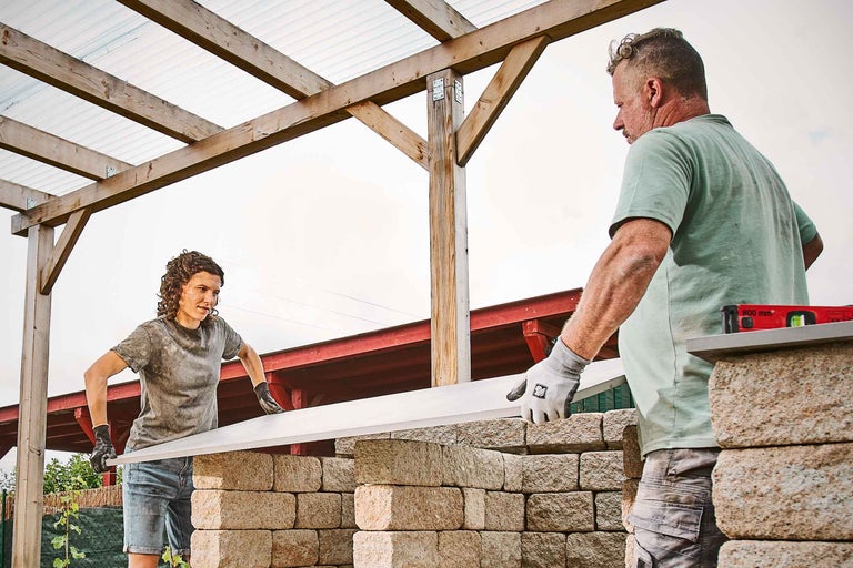 Deux personnes construisent un mur de jardin en blocs de béton sous un abri en bois.