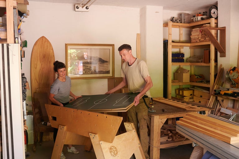 Deux personnes assemblent un baby-foot dans un atelier avec des étagères en bois et des outils.