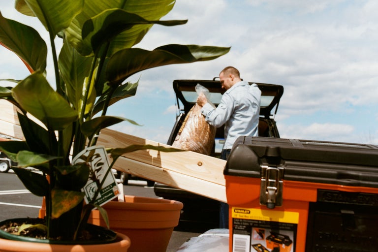 Szene mit Zimmerpflanze, Bauholz, Werkzeugkasten und Mann beim Ausladen von Baumaterial aus einem Auto.