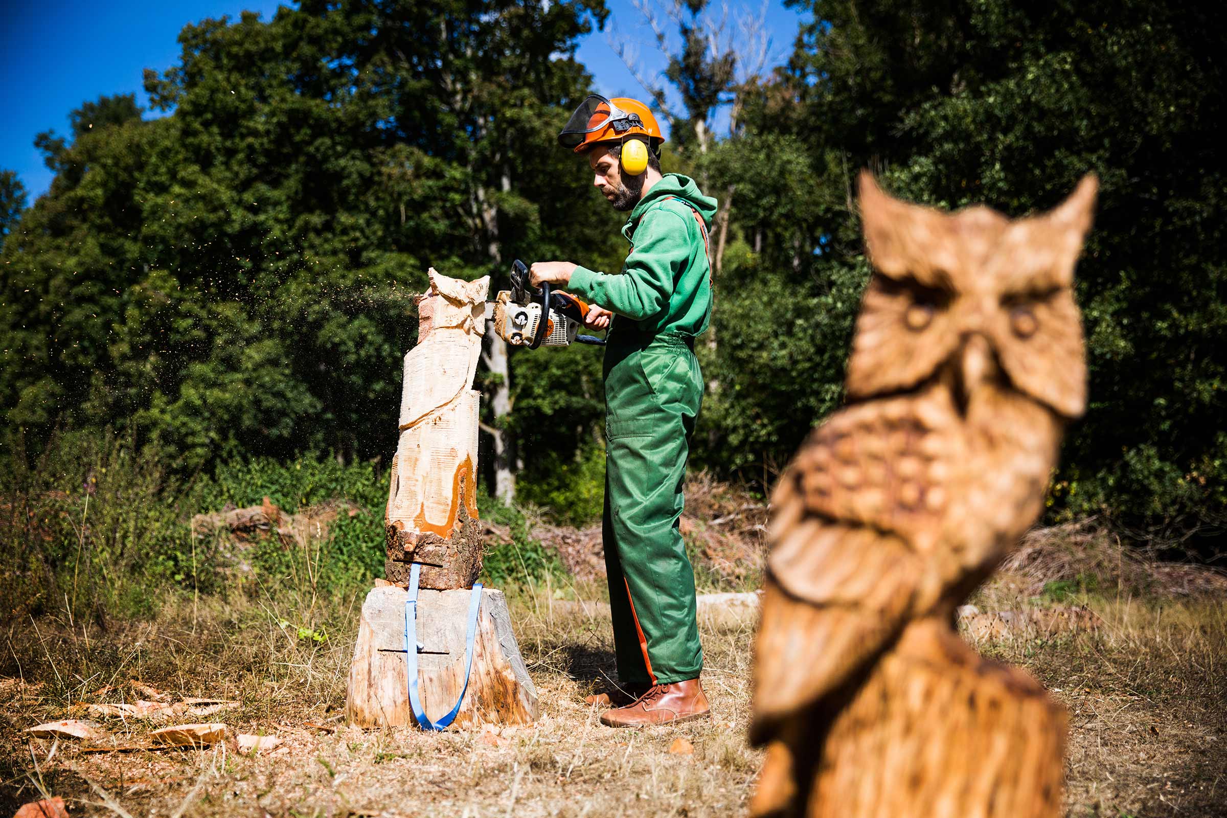 Un homme portant des équipements de protection sculpte une sculpture en bois à la tronçonneuse, avec une chouette sculptée au premier plan
