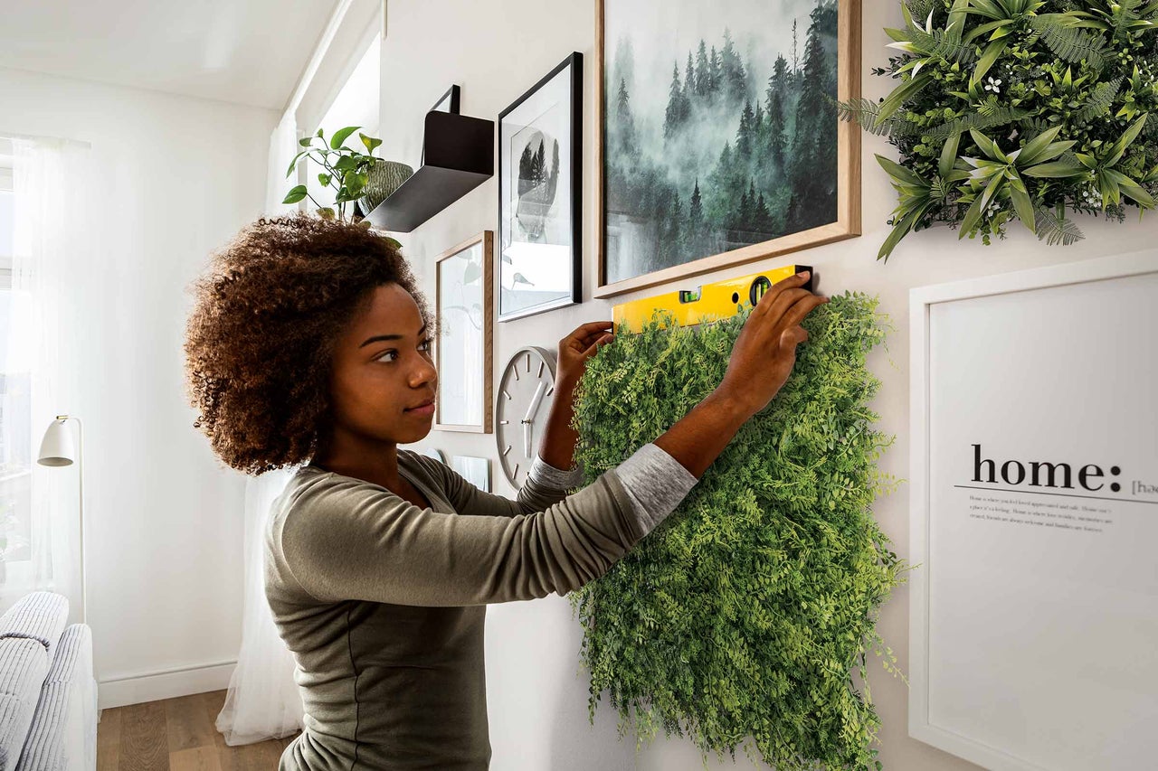 Frau montiert eine grüne Pflanzenwand mit einer Wasserwaage an einer Wand, umgeben von Dekorationen. Helle, organisierte Raumatmosphäre.