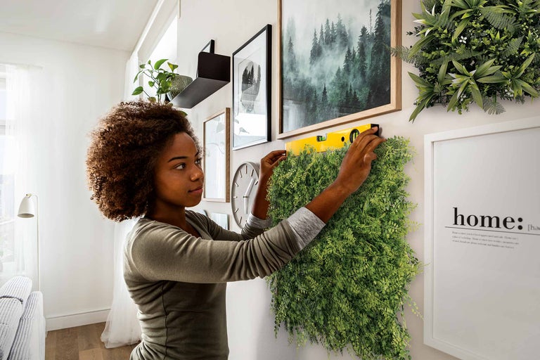 Frau montiert eine grüne Pflanzenwand mit einer Wasserwaage an einer Wand, umgeben von Dekorationen. Helle, organisierte Raumatmosphäre.