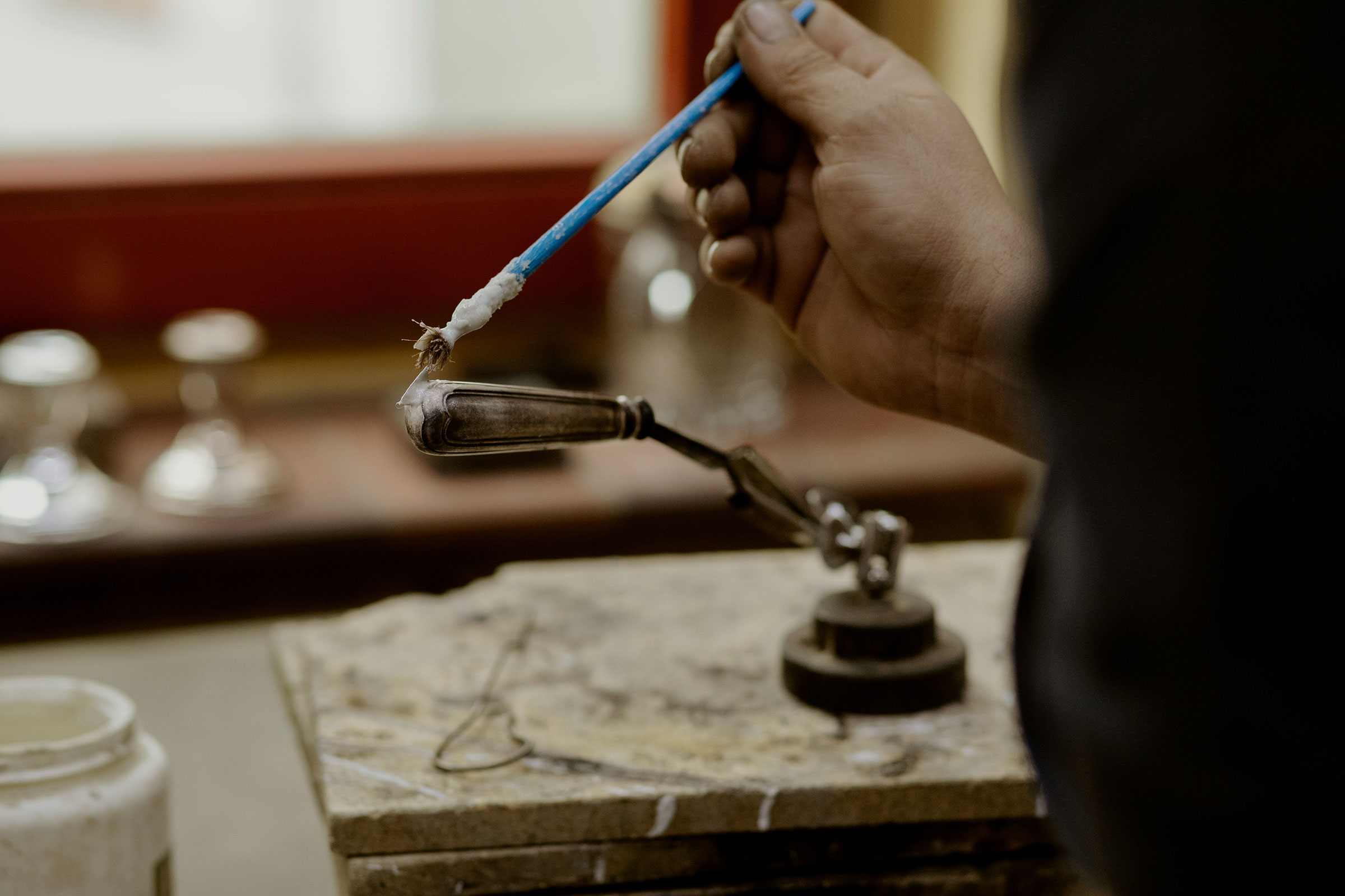Eine Hand hält einen Pinsel zum Auftragen von Flussmittel auf ein Metallstück, das in einem Halter befestigt ist. Werkstattszene mit Werkzeugen für die Metallbearbeitung.
