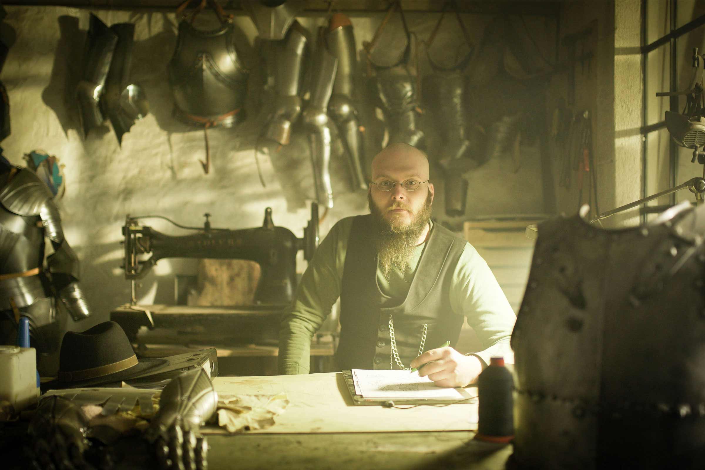 Atelier avec des pièces d'armure, un homme avec une barbe et des lunettes, une machine à coudre et un bureau avec un stylo et du papier.