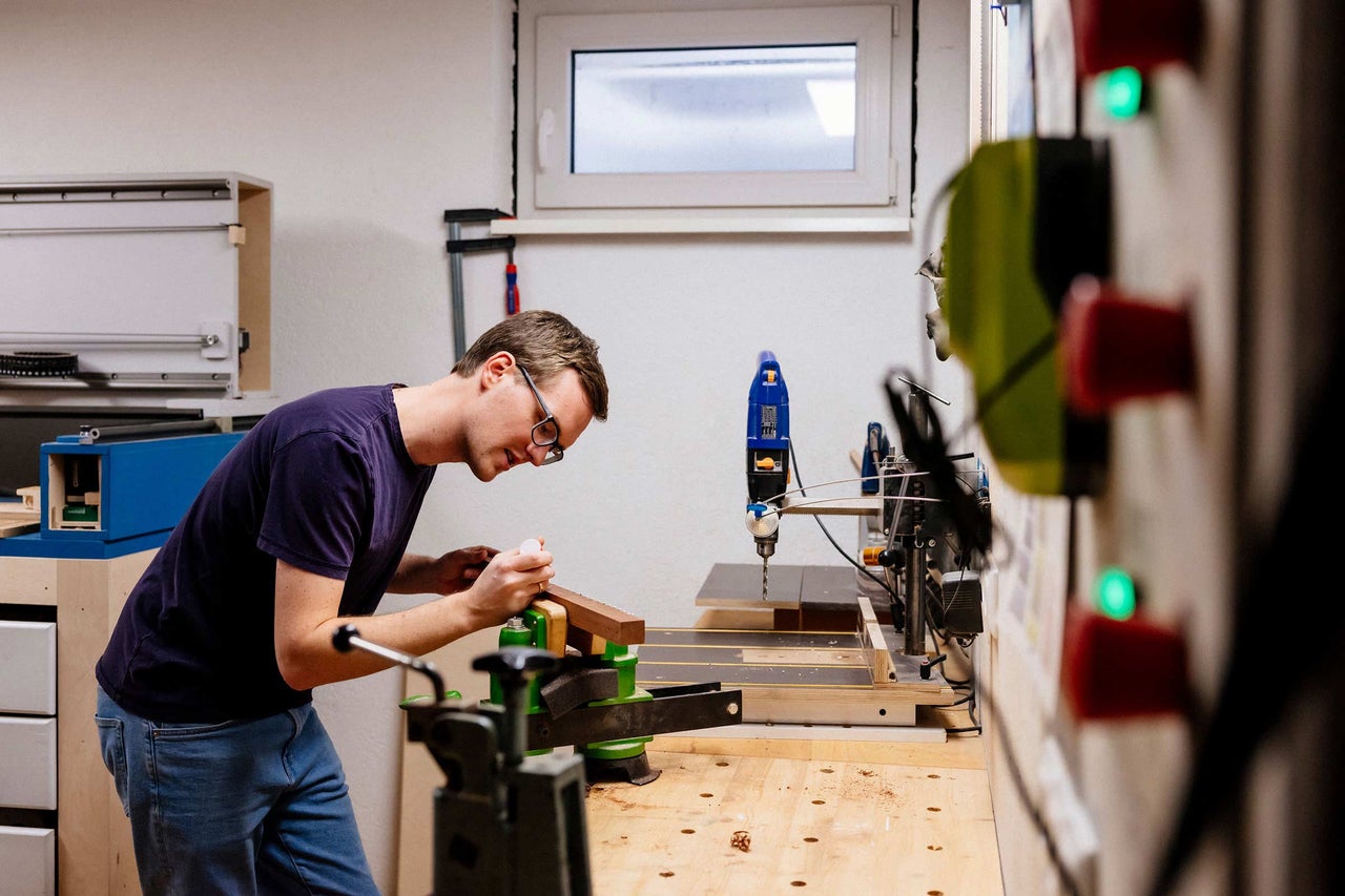 Un homme travaille sur une pièce de bois avec un établi, un étau et une perceuse à colonne dans un atelier.