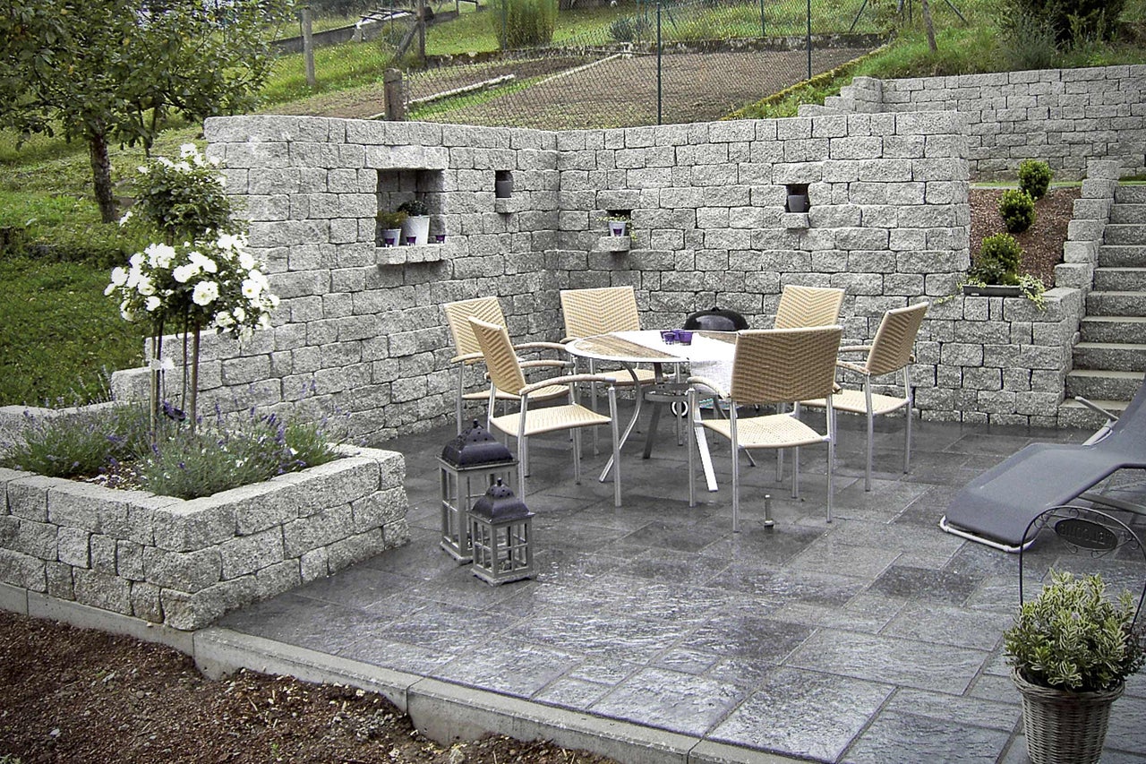 Gestalteter Gartenbereich mit Steinmauer, Pflastersteinen, Tisch, Stühlen und Pflanzen