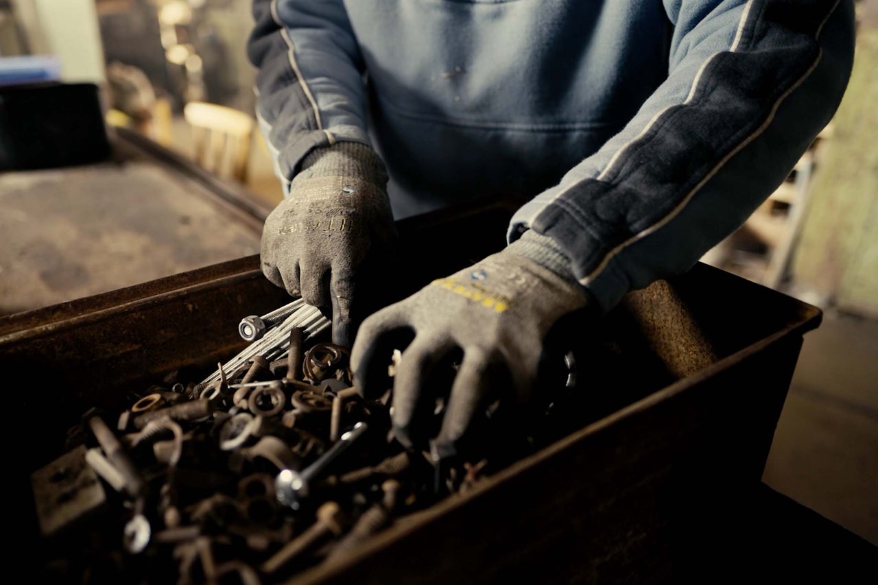 Une personne fouille une caisse de vis et d'écrous avec des gants de travail.