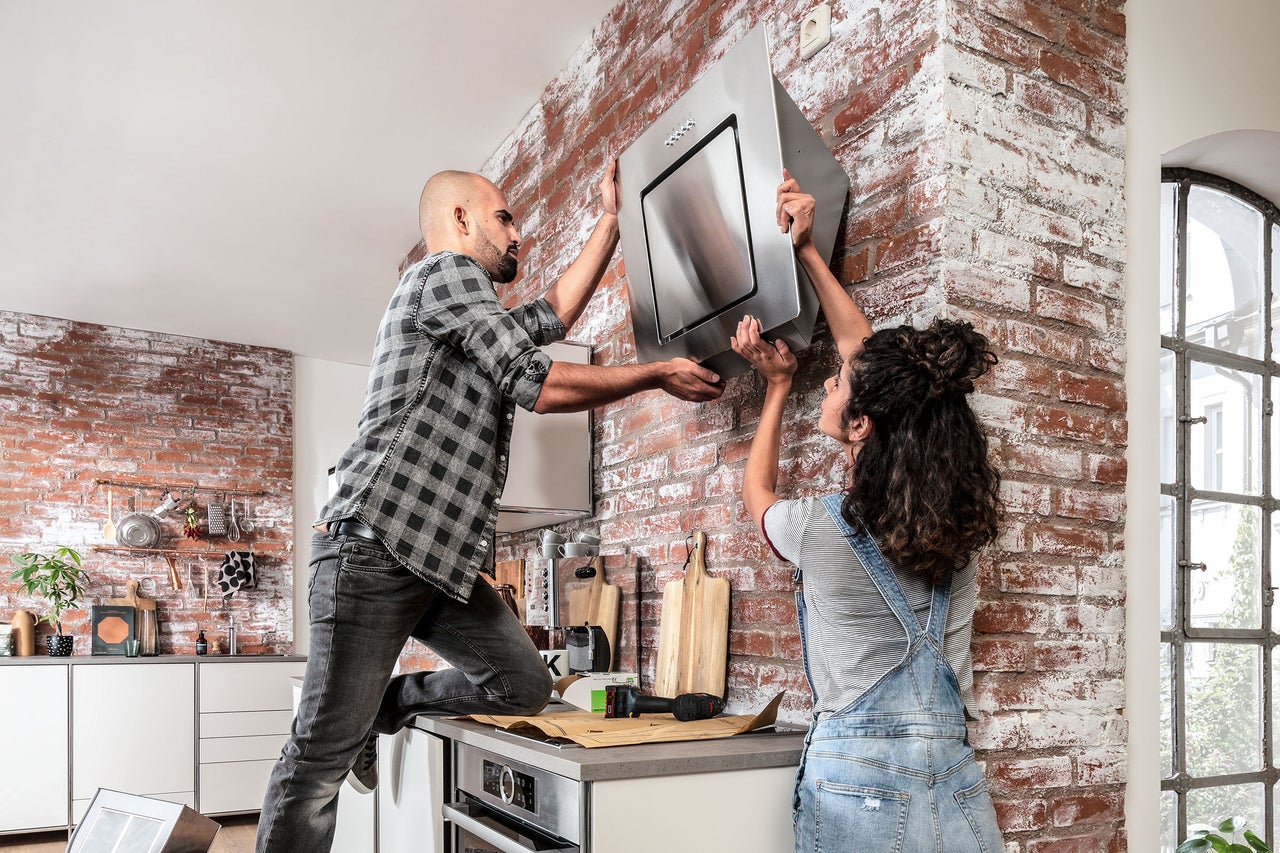 Zwei Personen installieren eine Dunstabzugshaube in einer Küche mit Ziegelwand