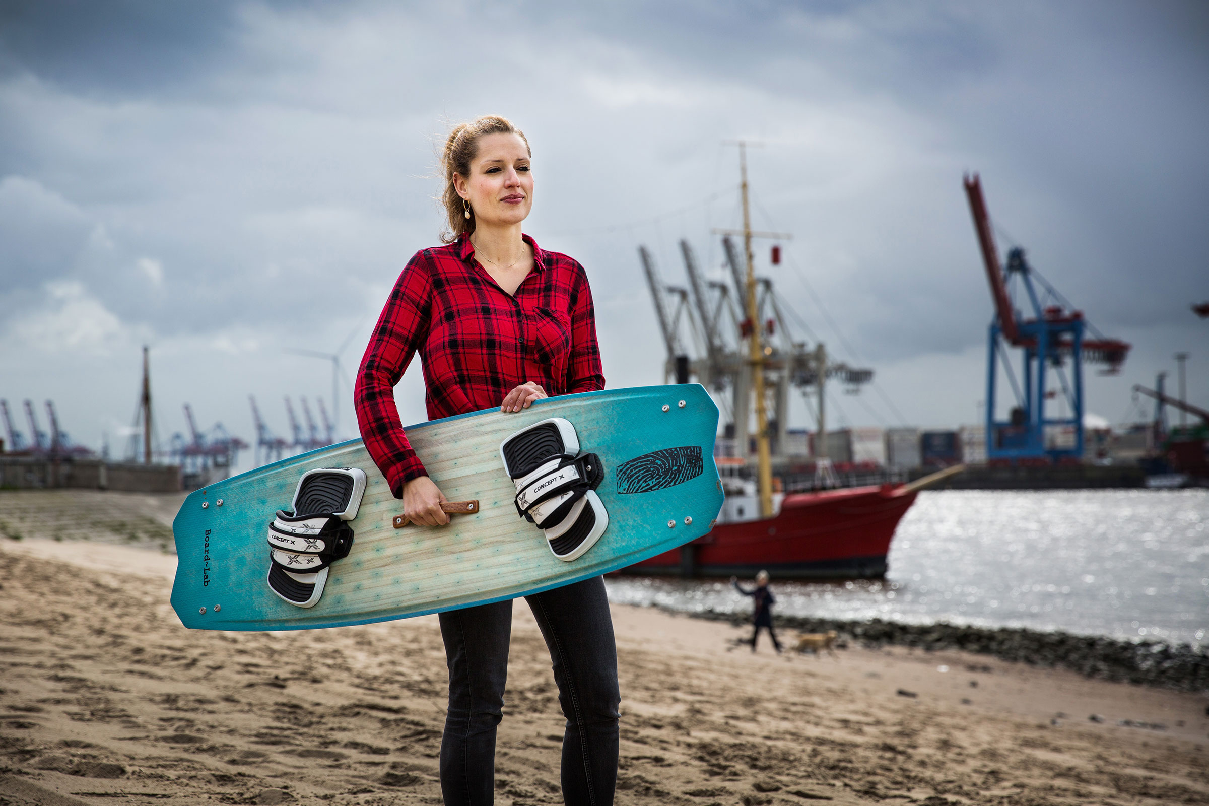 Femme tenant un kiteboard sur la plage avec des navires et des grues en arrière-plan