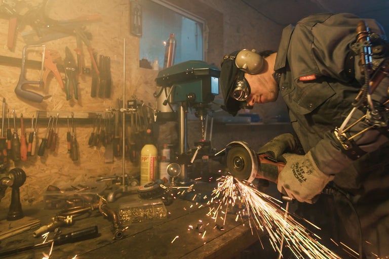 Ein Handwerker bearbeitet ein Metallstück mit einem Winkelschleifer, wobei Funken sprühen. Werkzeuge und eine Werkbank sind im Hintergrund zu sehen.