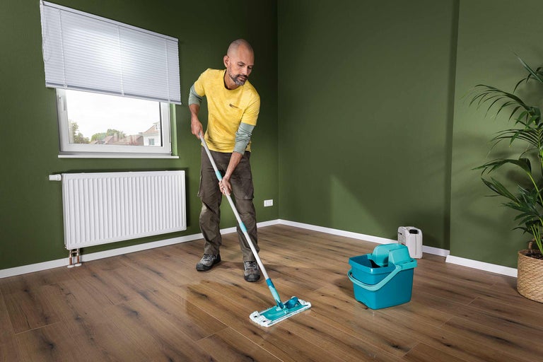 Ein Mann wischt mit einem Bodenwischer einen Holzboden in einem Raum mit Fenster, Heizkörper und Topfpflanze