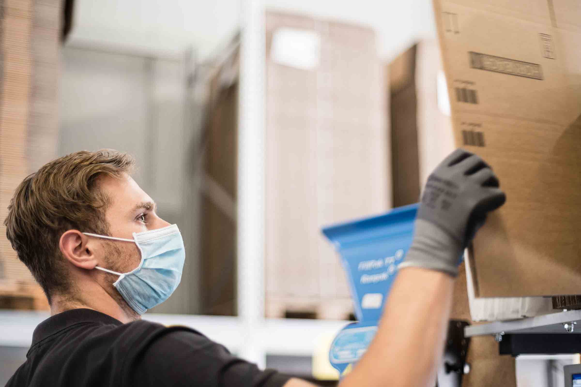 Un homme portant un masque de protection et des gants de travail manipule un carton dans un entrepôt.