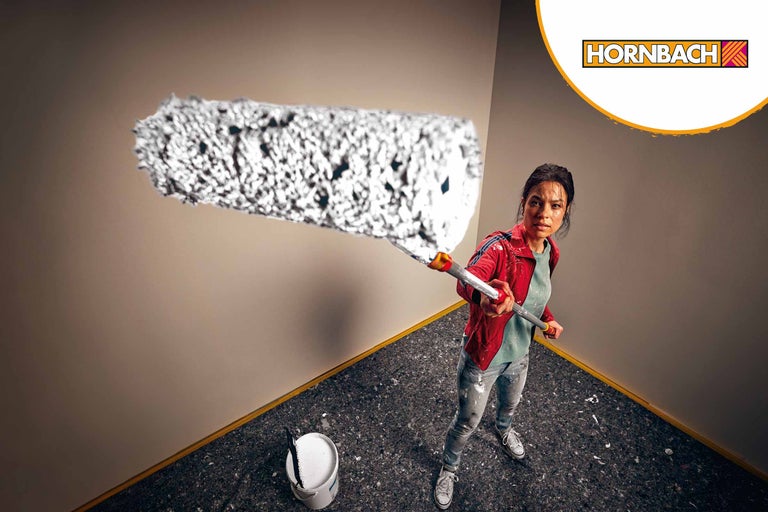 Une femme peint un mur avec un rouleau à peinture, un seau de peinture est dans la pièce, logo Hornbach