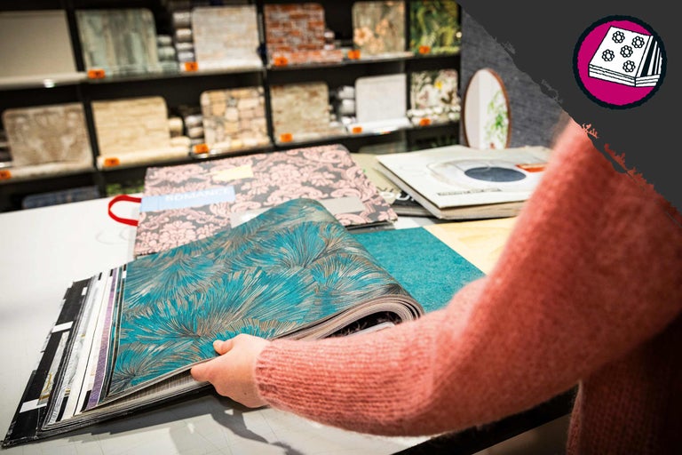 Eine Person blättert in einem Tapetenmusterbuch auf einem Tisch im Hornbach. Im Hintergrund befinden sich Regale mit weiteren Tapetenmustern.