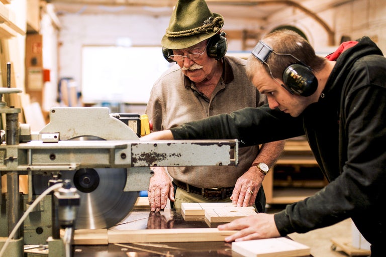 Zwei Personen mit Gehörschutz arbeiten an einer Tischkreissäge in einer Werkstatt.