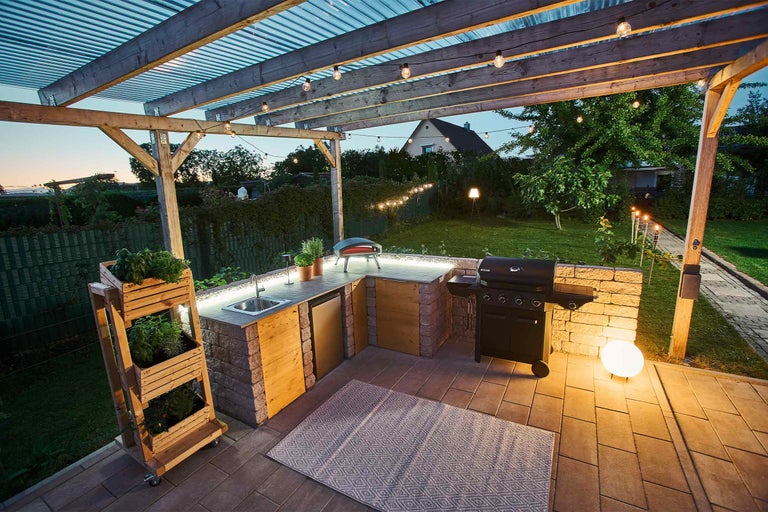 Cuisine d ete sous pergola en bois avec barbecue a gaz, meubles aspect pierre, portes en bois, evier et four a pizza sur terrasse pavee.