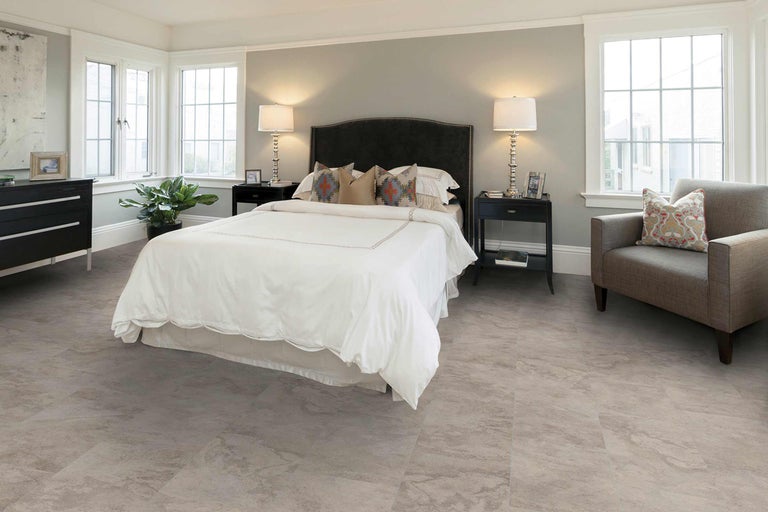 Chambre avec lit, tables de chevet, commode, fauteuil et revêtement de sol aspect pierre pour une ambiance claire et conviviale.