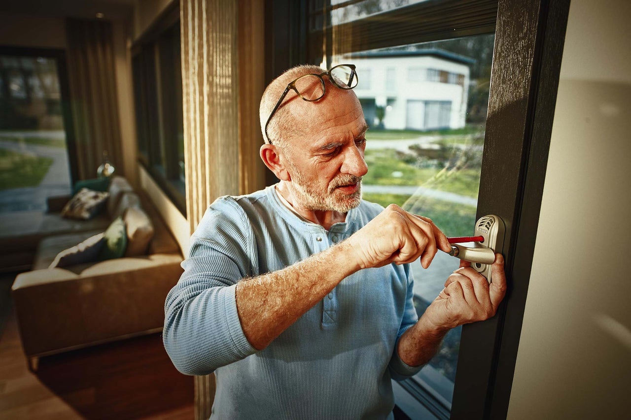 Ein Mann montiert einen abschließbaren Fenstergriff mit einem Schraubendreher an einem modernen Fenster in einem Wohnraum.