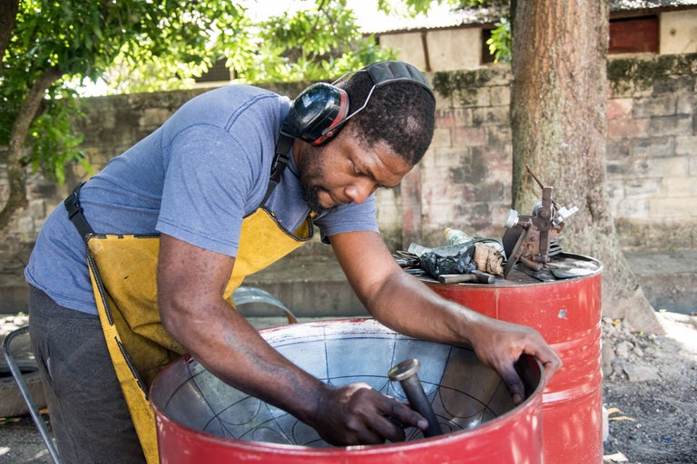Ein Mann mit Ohrenschützern formt eine Steeldrum mit einem Hammer. Er trägt eine Lederschürze.