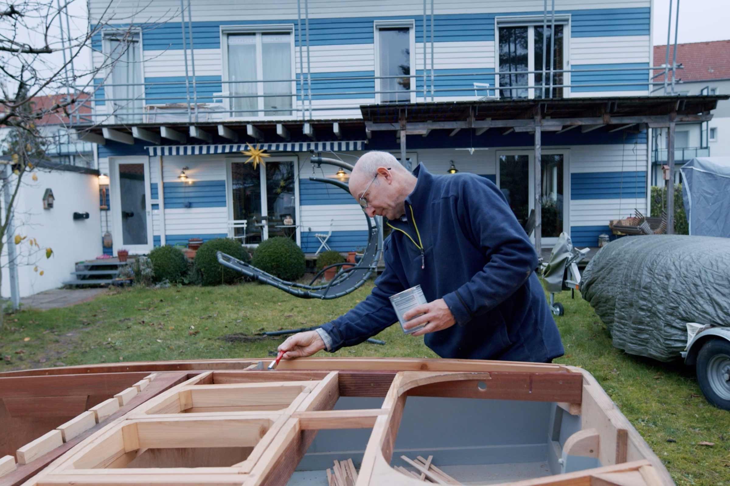 Un homme peint un bateau en bois avec un pinceau et une boîte de peinture dans le jardin d'une maison.