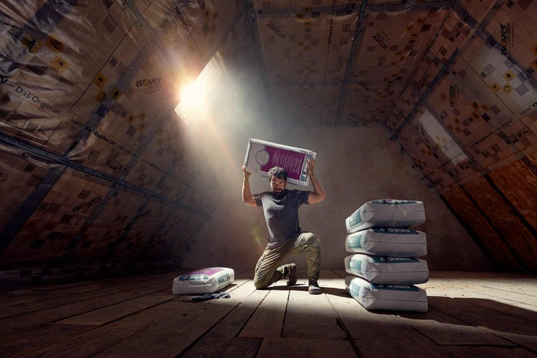 Ein Mann kniet auf einem Dachboden und hält einen Sack Estrichmörtel über dem Kopf. Im Hintergrund sind weitere Säcke Estrichmörtel und eine Dachisolierung zu sehen.