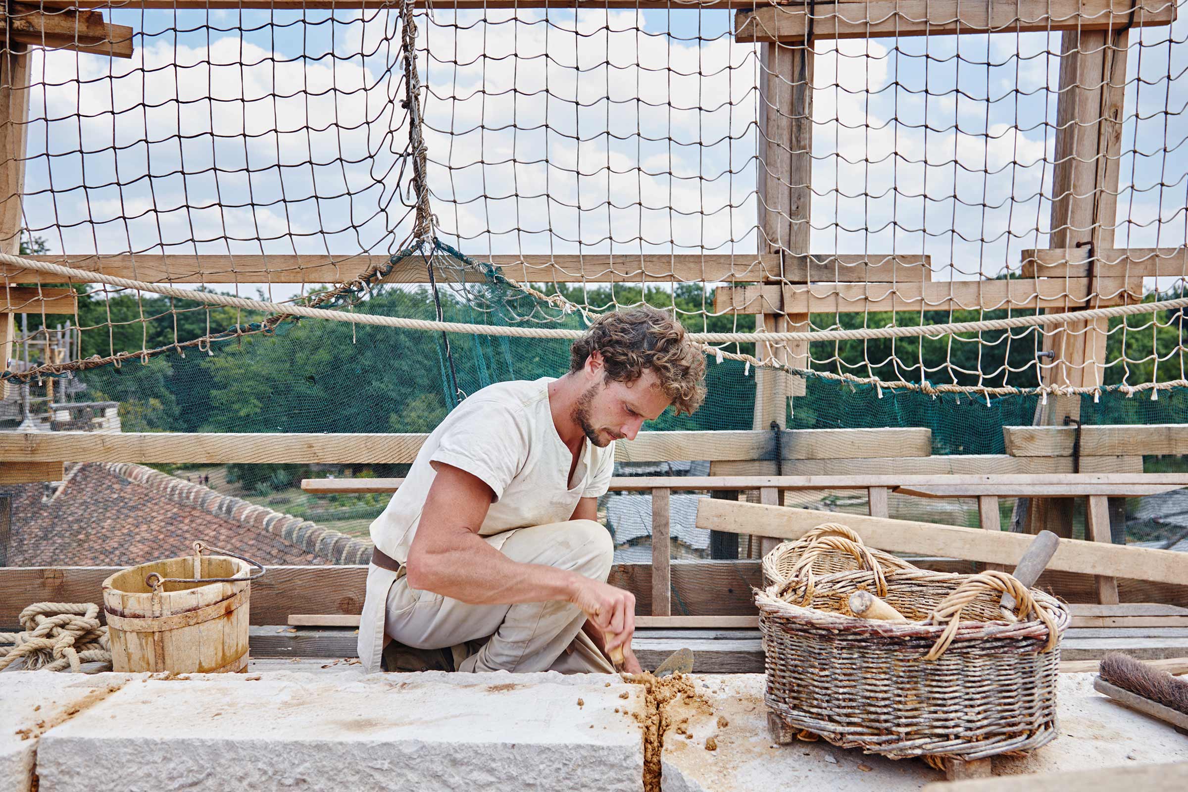 Un artisan est agenouillé sur un chantier et travaille la pierre avec des outils. Un seau en bois et un panier en osier sont visibles à l'arrière-plan.