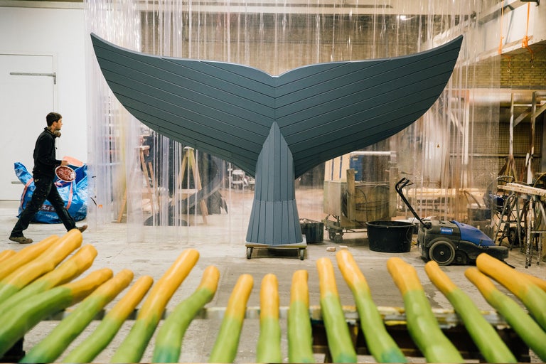 Scène d'atelier avec une grande queue de baleine en bois sur un chariot à roulettes et des piquets en bois peints au premier plan