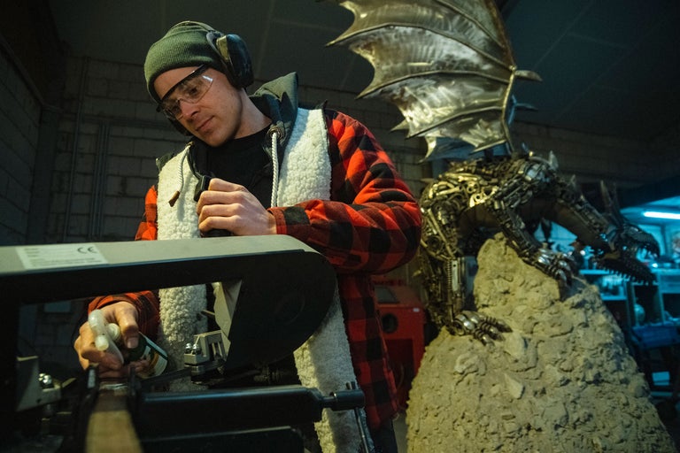 Homme portant des lunettes de sécurité et une protection auditive manipulant une machine, avec une sculpture de dragon en métal en arrière-plan.