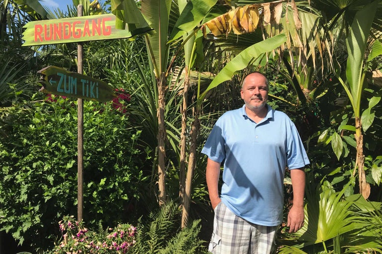 Un homme se tient à côté d'un panneau indiquant un circuit et la zone Tiki au milieu de plantes tropicales