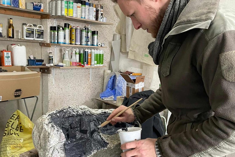 Un homme peint un rocher avec un pinceau et de la peinture dans un atelier, étagères avec de la peinture en arrière-plan.