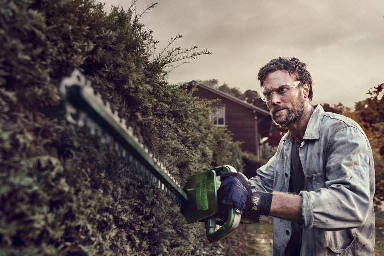 Mann schneidet Hecke mit Heckenschere, Schutzbrille und Handschuhen.