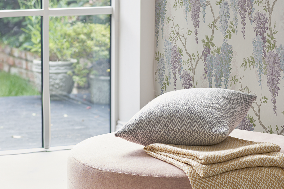 Wohnzimmer mit Vliestapete mit Blauregen Motiv, runder Sitzhocker mit Dekokissen und Wohndecke vor einem bodentiefen Fenster mit Blick in den Garten