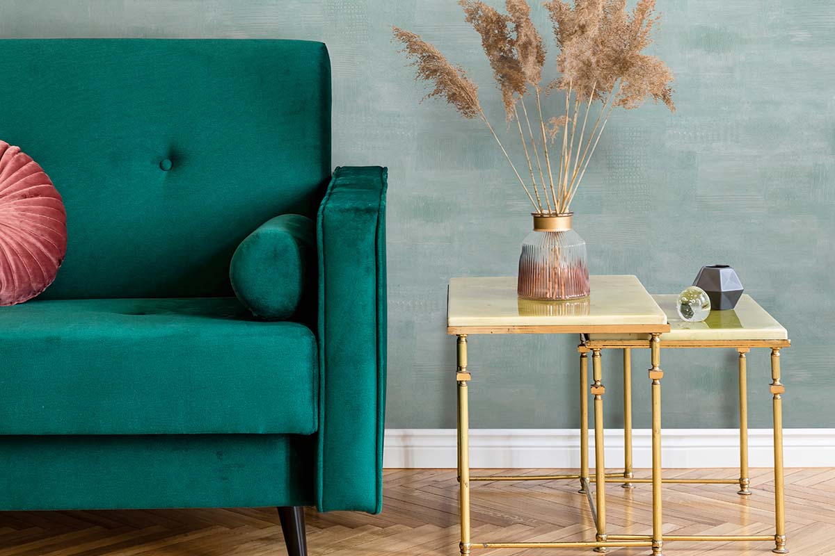 Wohnzimmer mit grünem Samtsofa, zwei Beistelltischen mit Metallgestell und Glasvase mit Trockenblumen vor einer strukturierten Wand.
