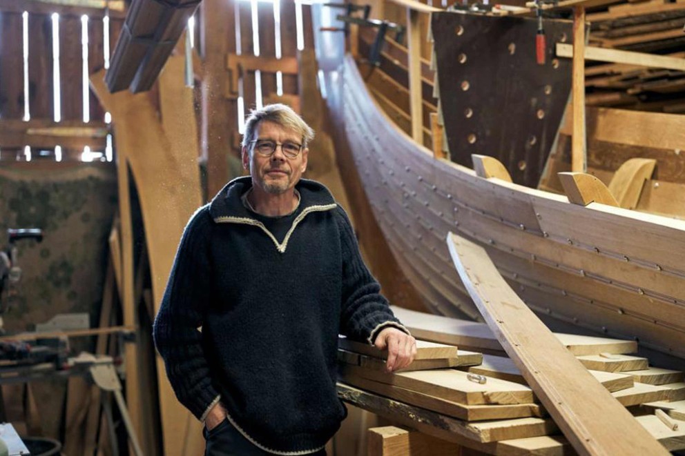 Un homme se tient dans un atelier devant un bateau en bois en construction et des poutres en bois.