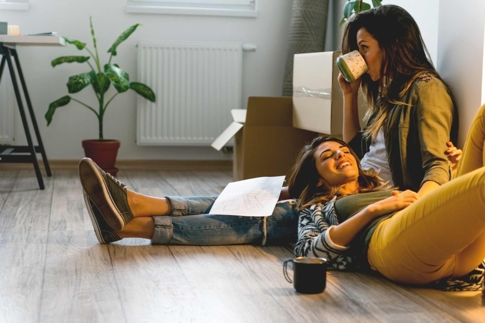 Deux femmes se détendent dans une pièce avec des cartons de déménagement, une plante d'intérieur et un radiateur.