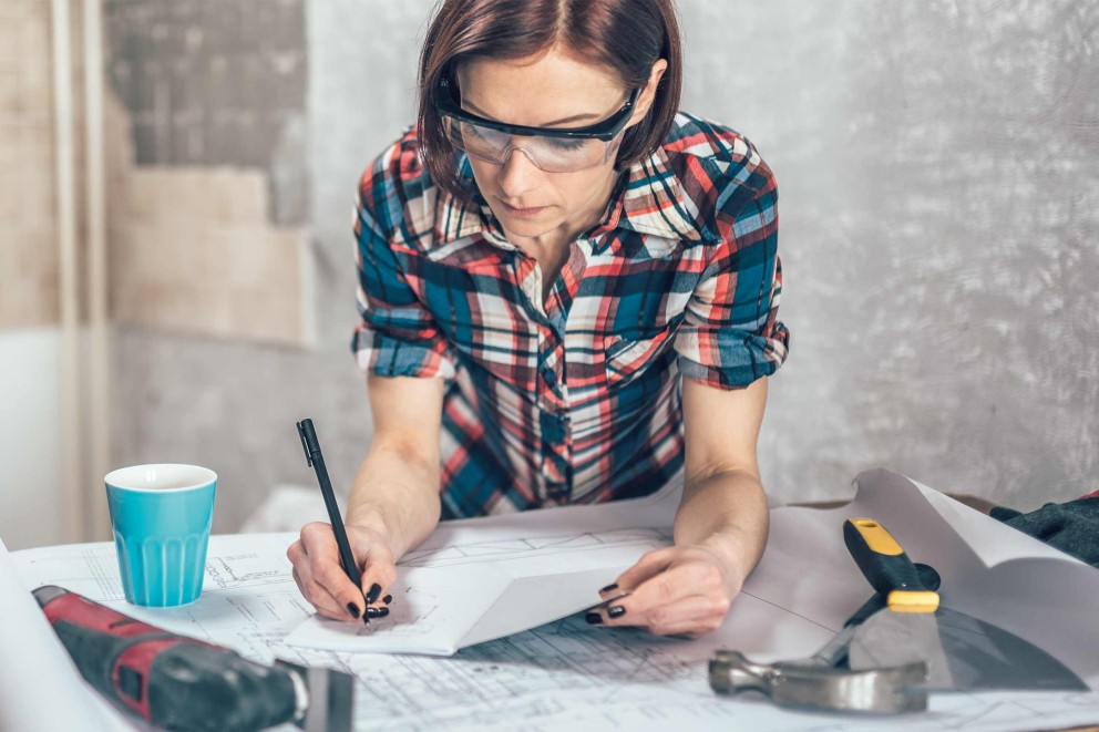 Frau mit Schutzbrille, Stift und Bauplan bei der Arbeit