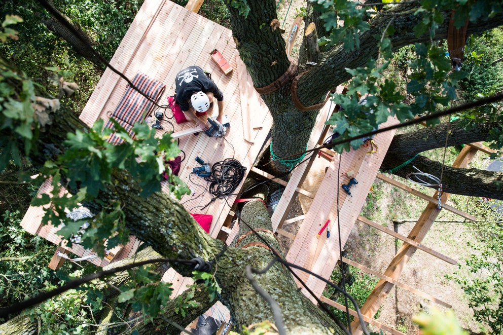 
				Pour Lucas Wahl, construire sa cabane dans les arbres à environ dix mètres de hauteur ne lui pose aucun problème. Ici, il fixe les planches sur la base de la cabane, tout en étant bien sûr assuré par les cordes.

			