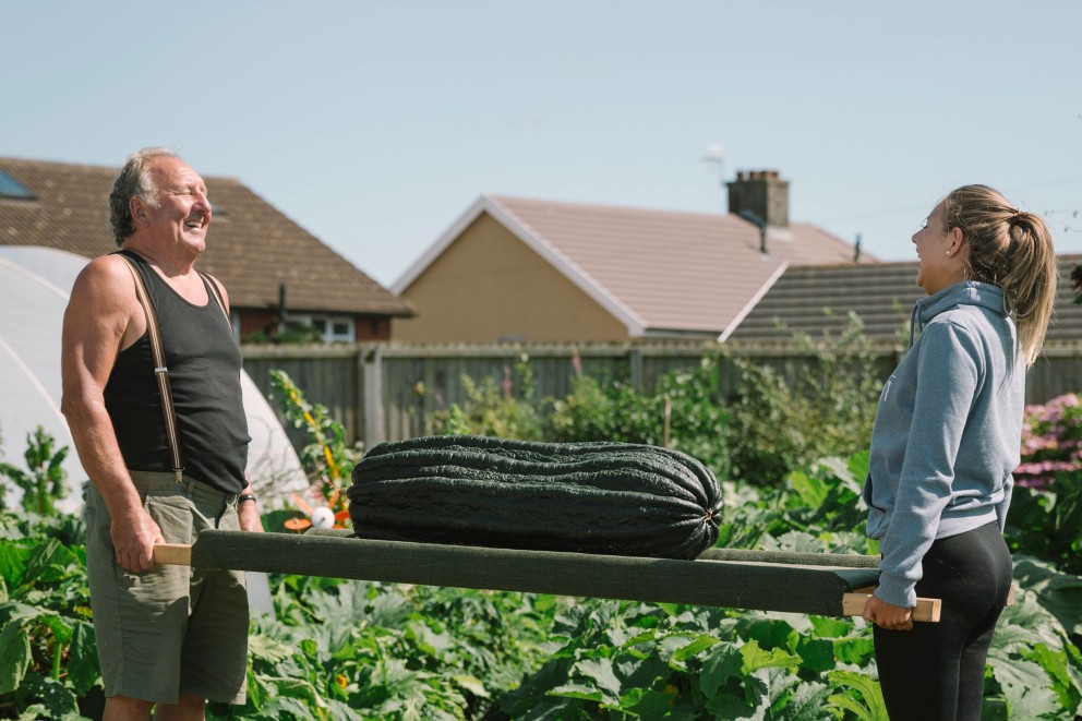 Deux personnes transportent une longue courge vert foncé sur une civière dans un jardin.