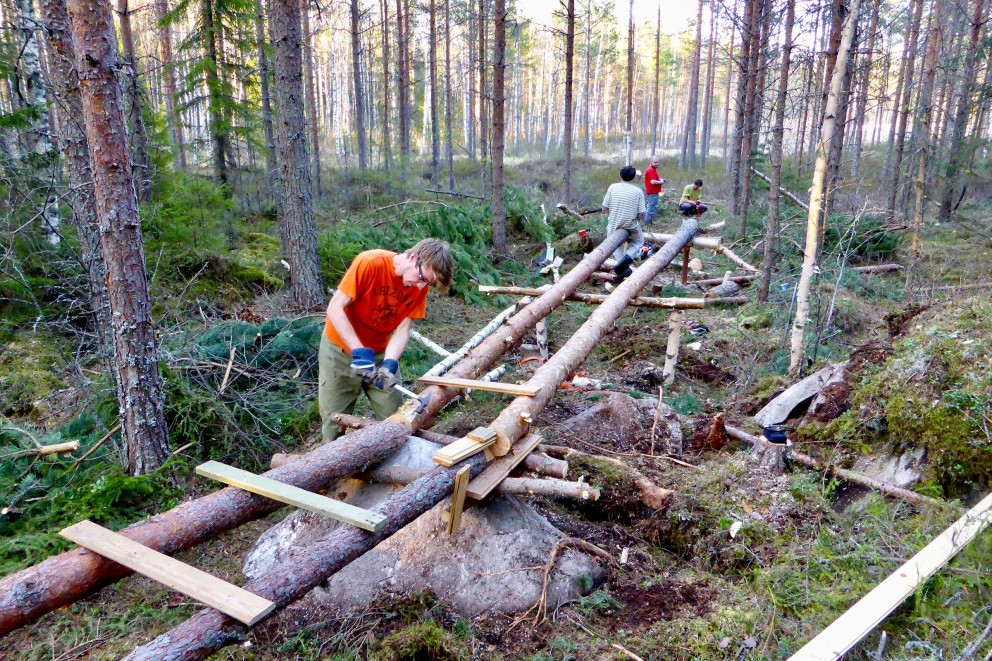 
				Das Sumpfgebiet in Finnland muss erstmal begehbar gemacht werden mithilfe eines Stegs. Die Unterkonstruktion setzt sich zusammen aus Kieferstämmen, die bis zu acht Meter tief in den Sumpfboden gerammt werden.

			